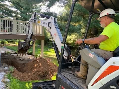 Worker operating a white excavator beside a house, digging a trench near a deck