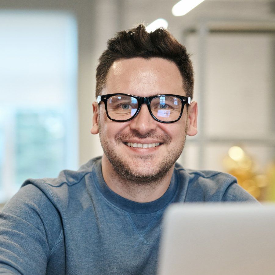 Smiling person in glasses at a laptop, wearing a blue sweater in a bright office setting