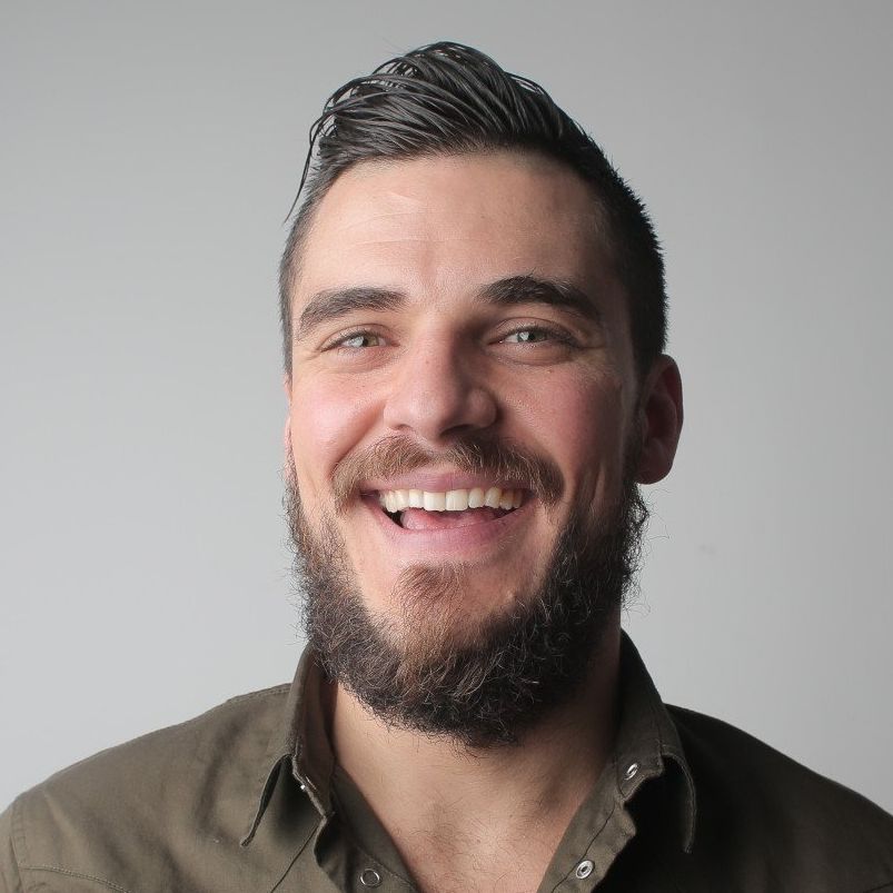 Smiling bearded man with dark hair in an olive shirt against a gray background