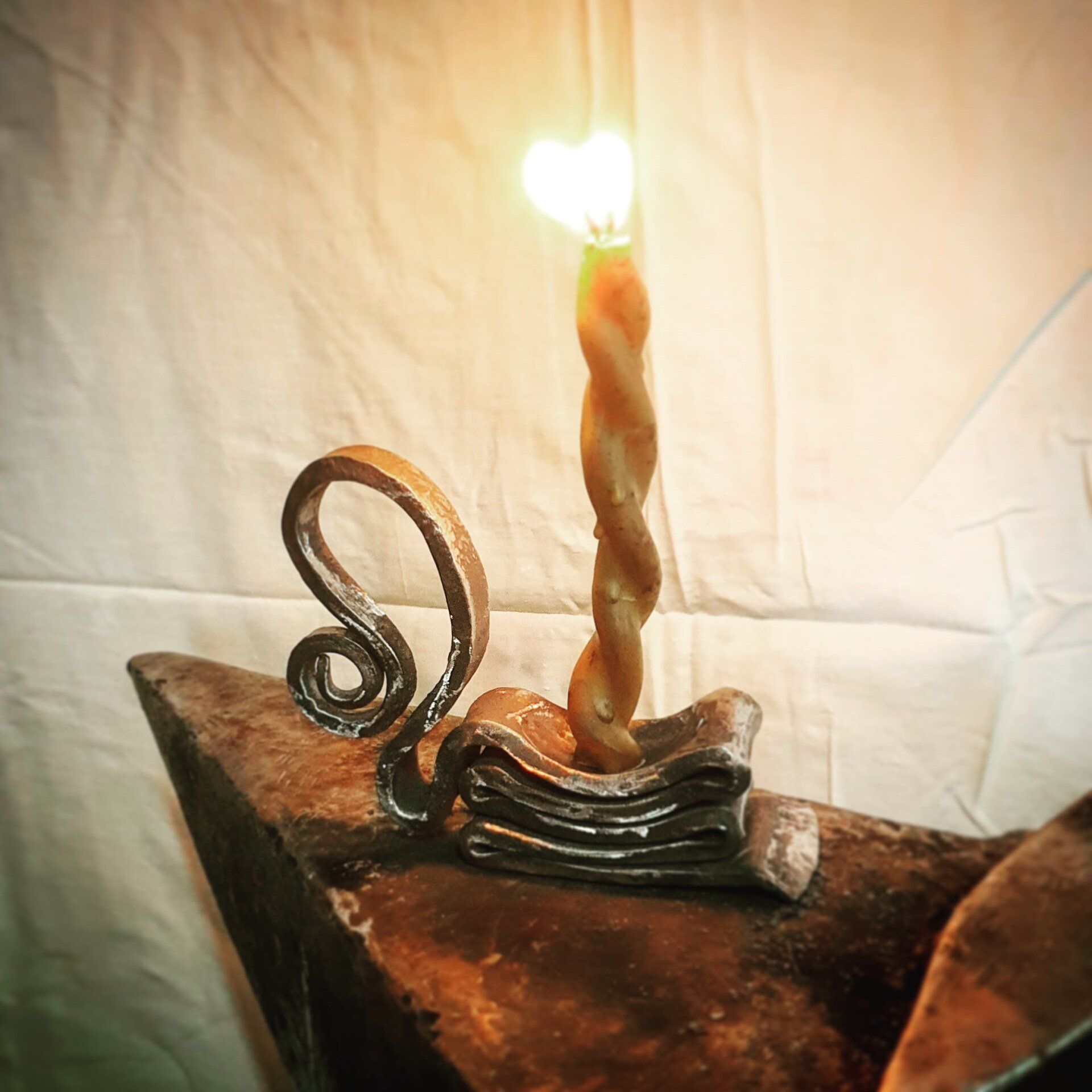Candela attorcigliata che brucia su una base decorativa scura accanto a una spirale di filo metallico, su uno sfondo di tessuto beige.