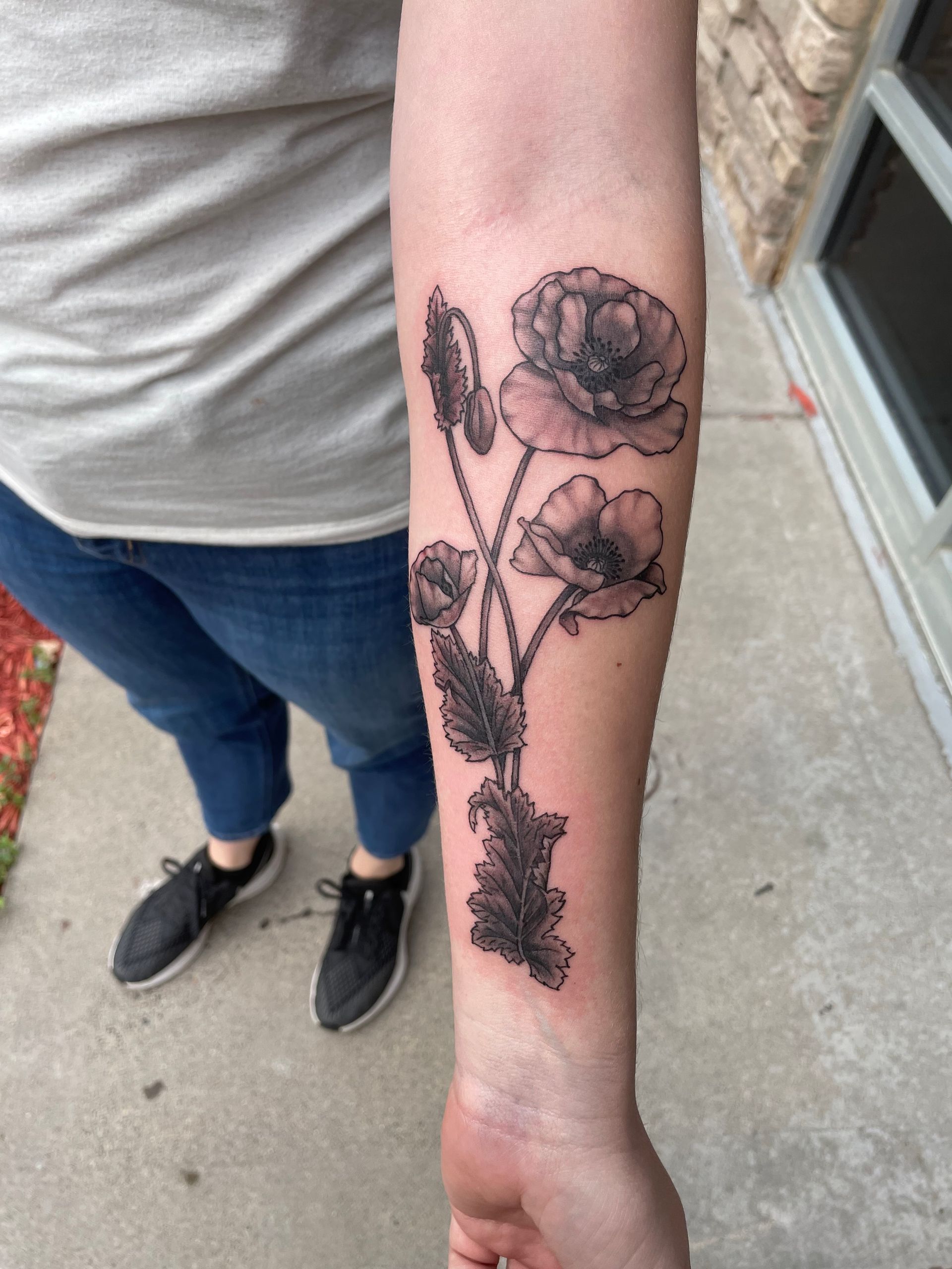Arm with a black and white poppy flower tattoo. Person wearing jeans and sneakers.