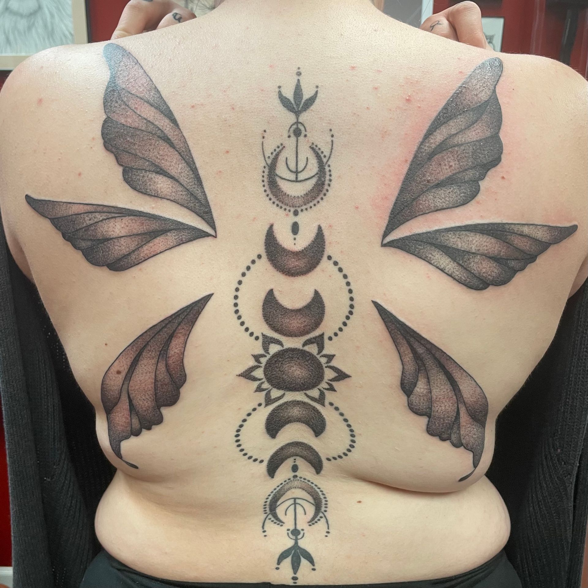 Back tattoo of fairy wings and a spine-centered moon phase design. The tattoo is gray and brown.