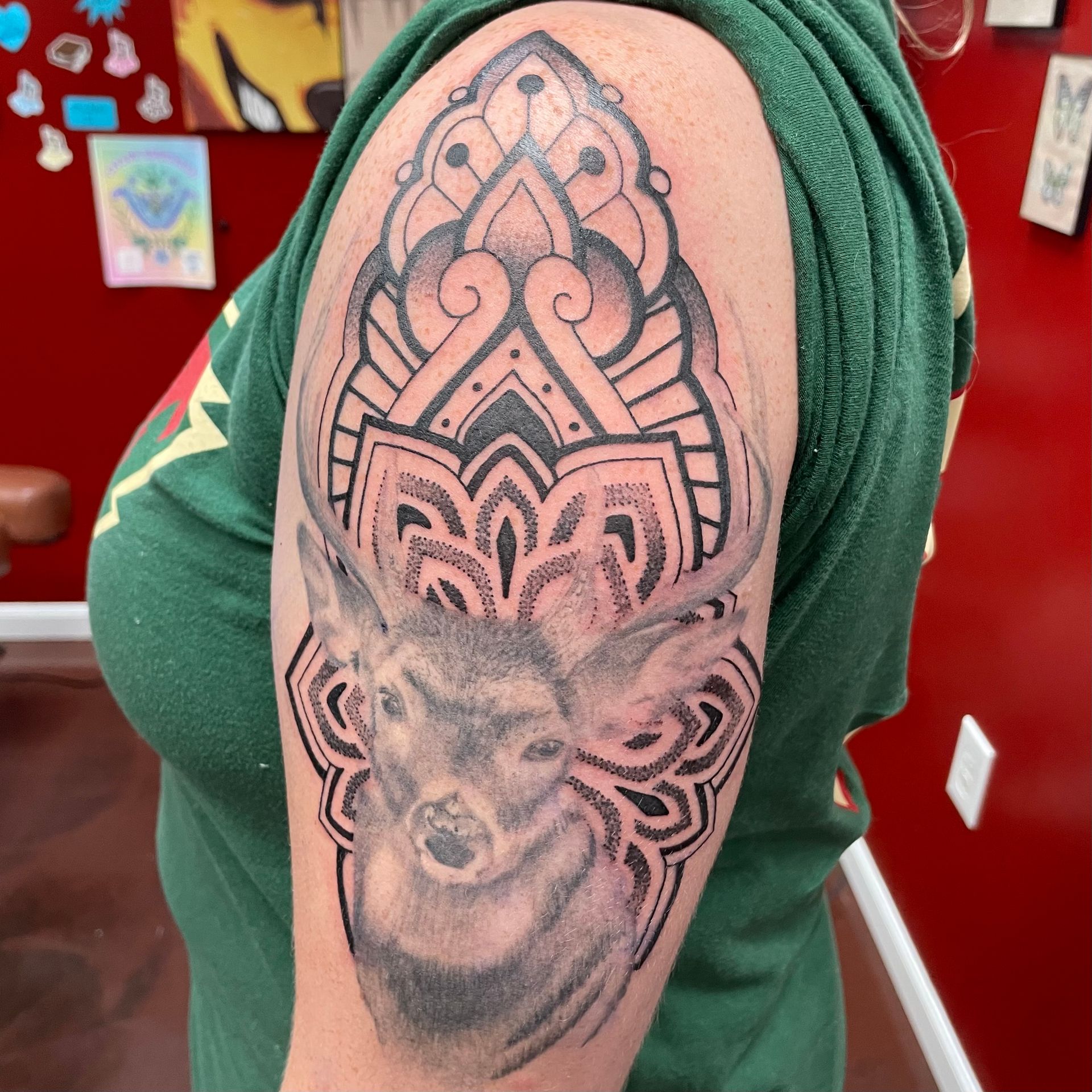Tattoo of a deer's head with a mandala design above it on a person's upper arm. The person wears a green shirt.