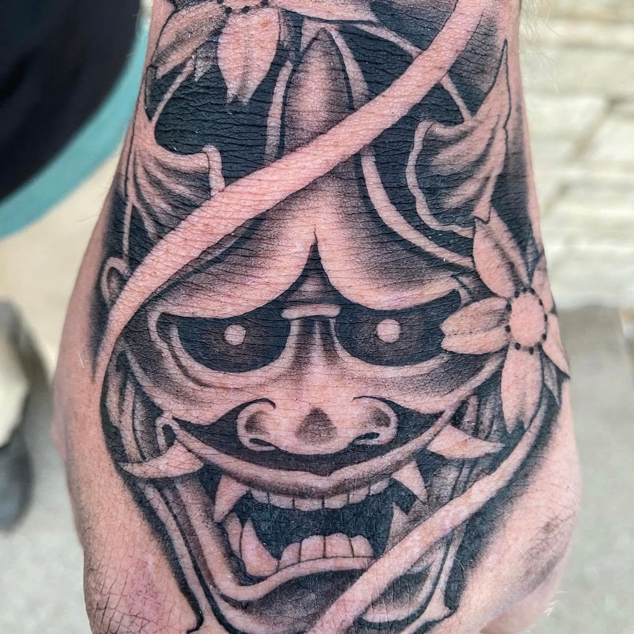 Black and grey Hannya mask tattoo on a hand, featuring sharp teeth, floral accents, and intricate shading.