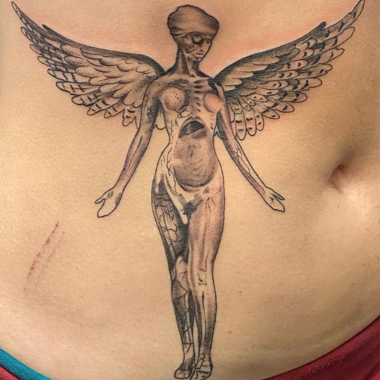 Tattoo of a nude figure with angel wings, arms outstretched. Black and gray ink. On a person's torso.