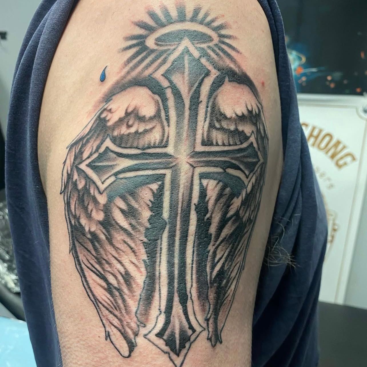 Tattoo on upper arm: black cross with angel wings, halo, and radiating beams.