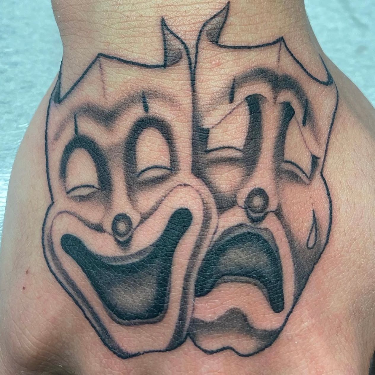 Tattoo of comedy and tragedy masks on a hand, black ink with shading.