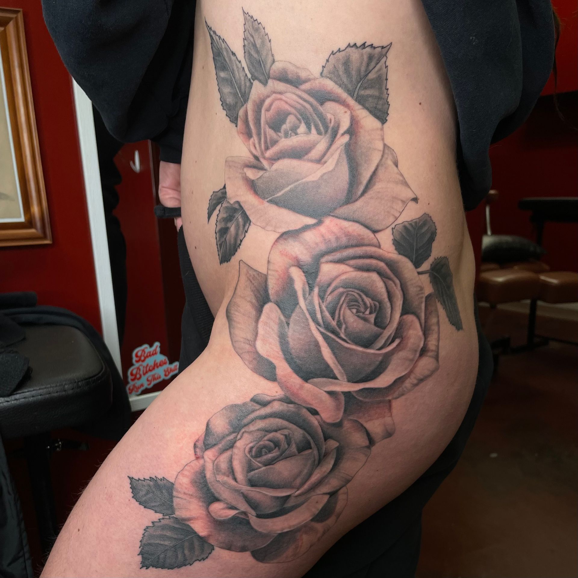 Tattoo of three grey roses and leaves, extending down a person's thigh.