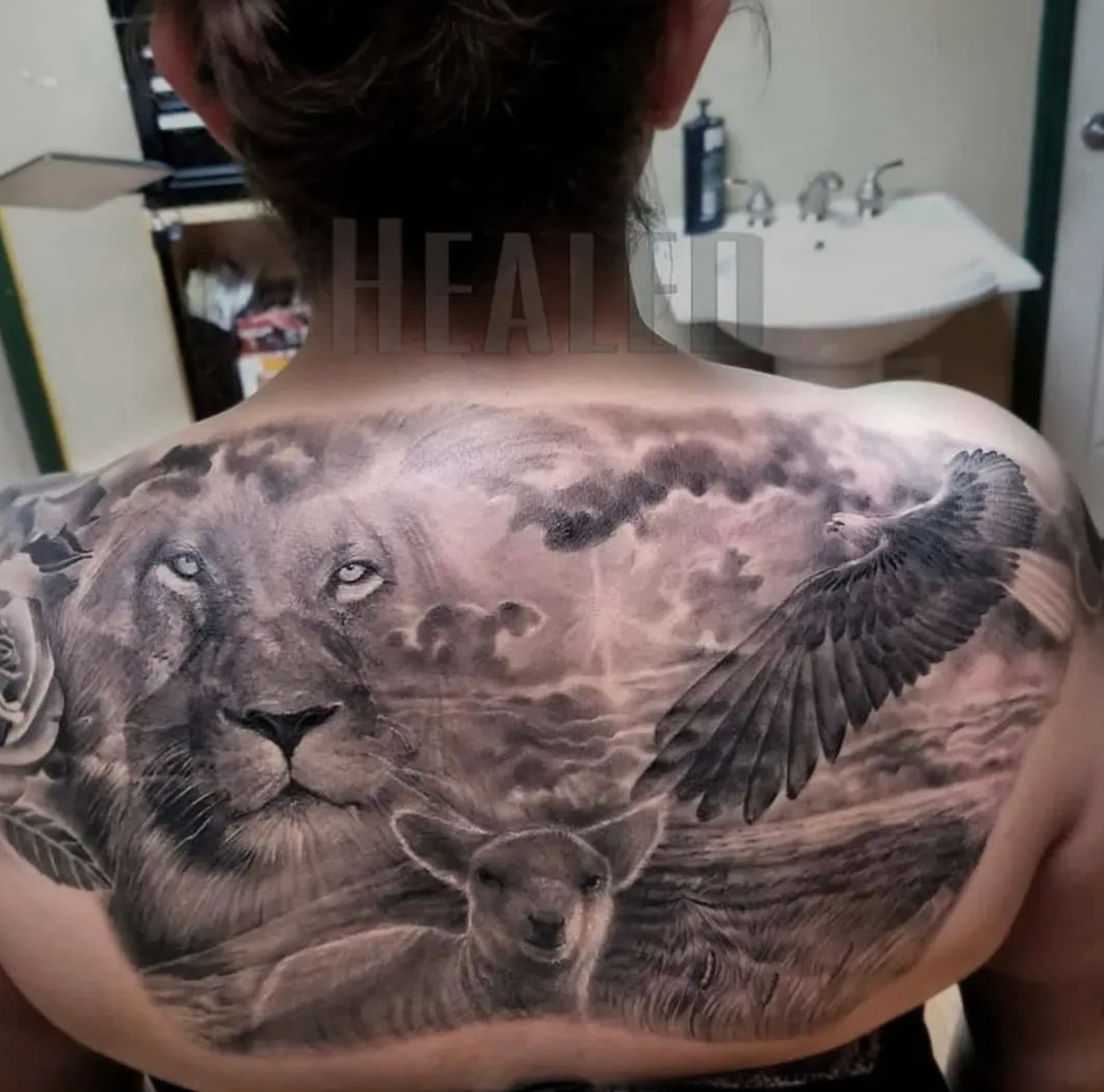 Tattoo on a back depicts a lion, eagle, lamb, and rose against a cloudy sky background.