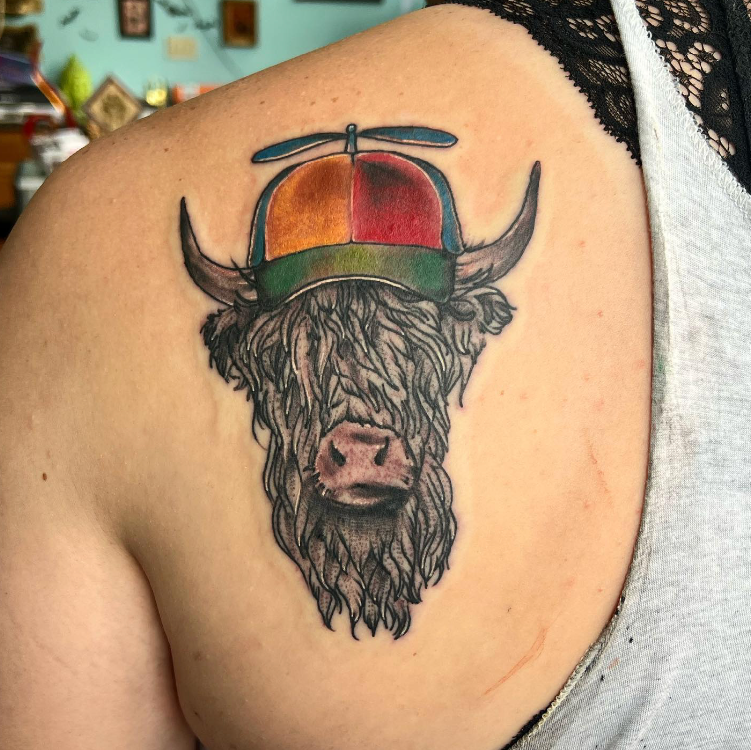Tattoo of a Highland cow wearing a colorful hat with a propeller on a person's shoulder.