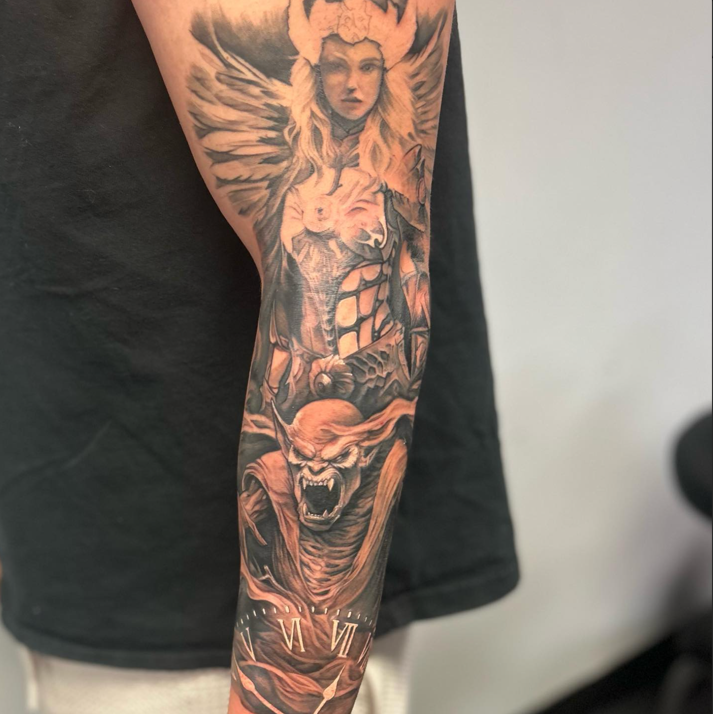 Tattoo on arm, showing angel-like figure above a demon-like creature; black and gray shading.
