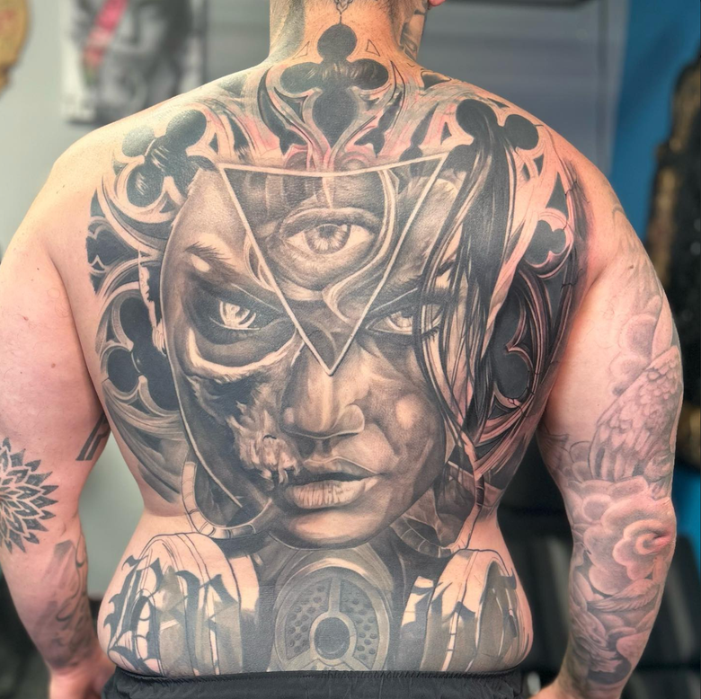 Tattoo on a person's back, a gothic-style face with an eye within a triangle. Detailed and grayscale.