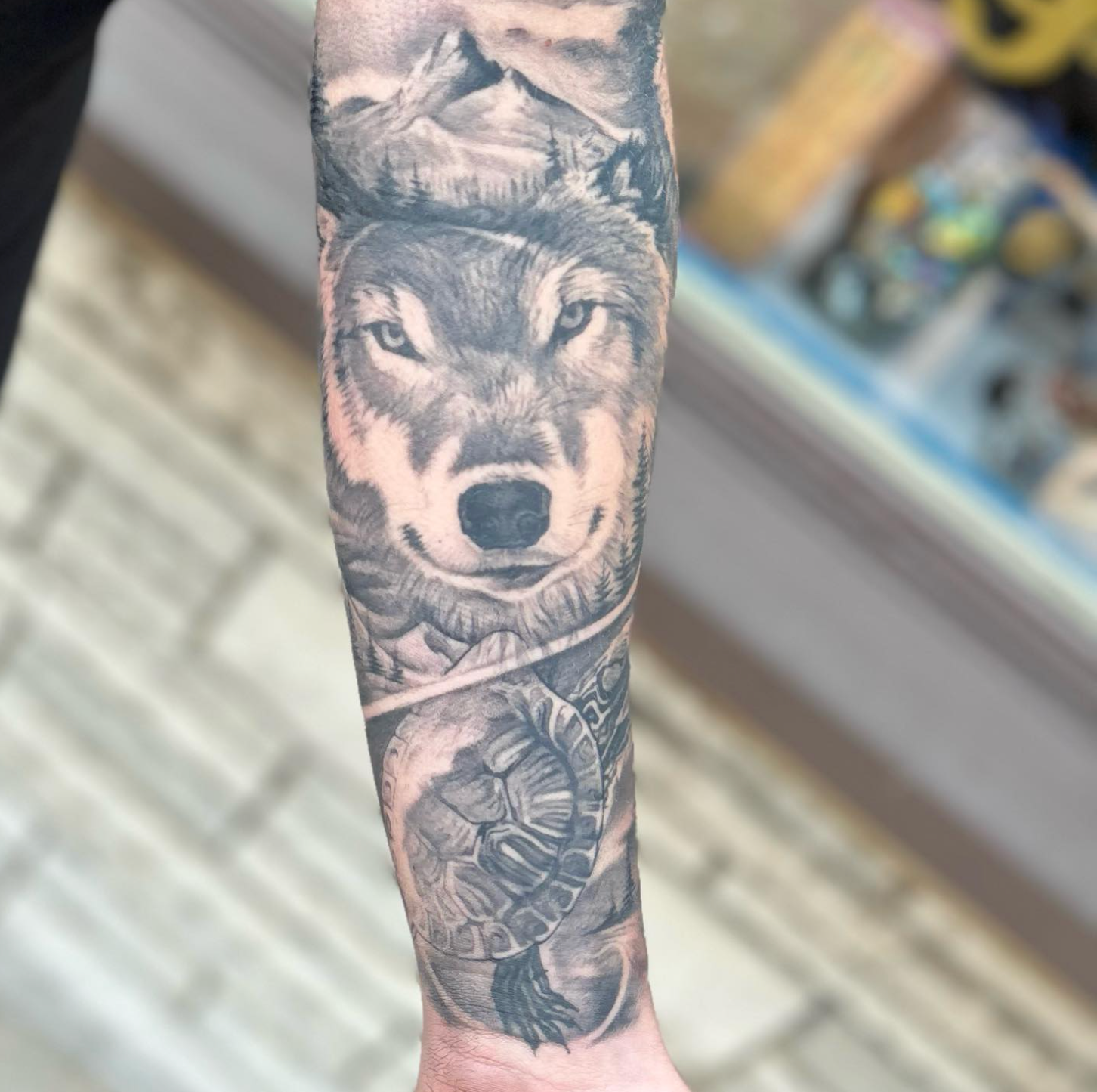 Black and gray forearm tattoo of a wolf with mountain backdrop and turtle below.