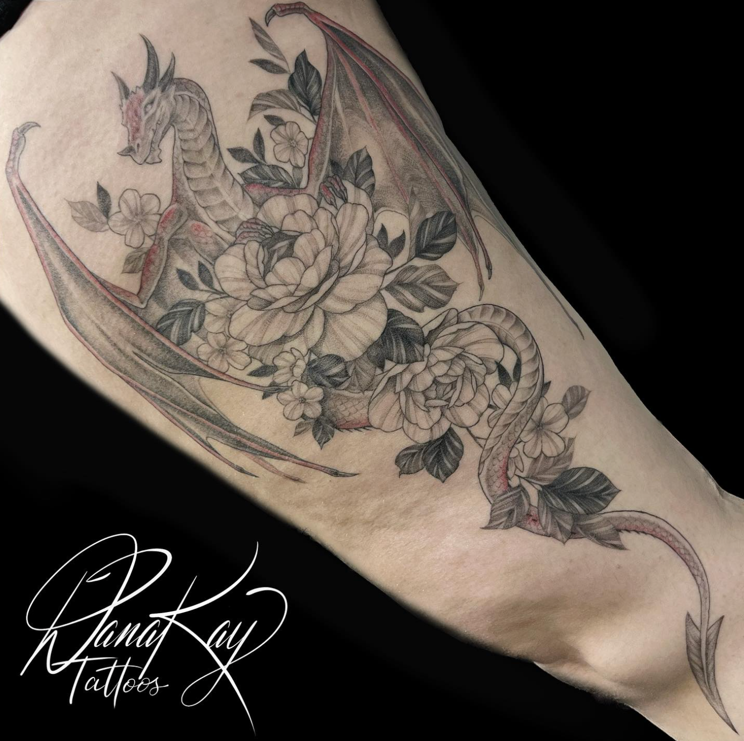Tattoo of a dragon with large wings and flowers, black and grey ink on an arm.