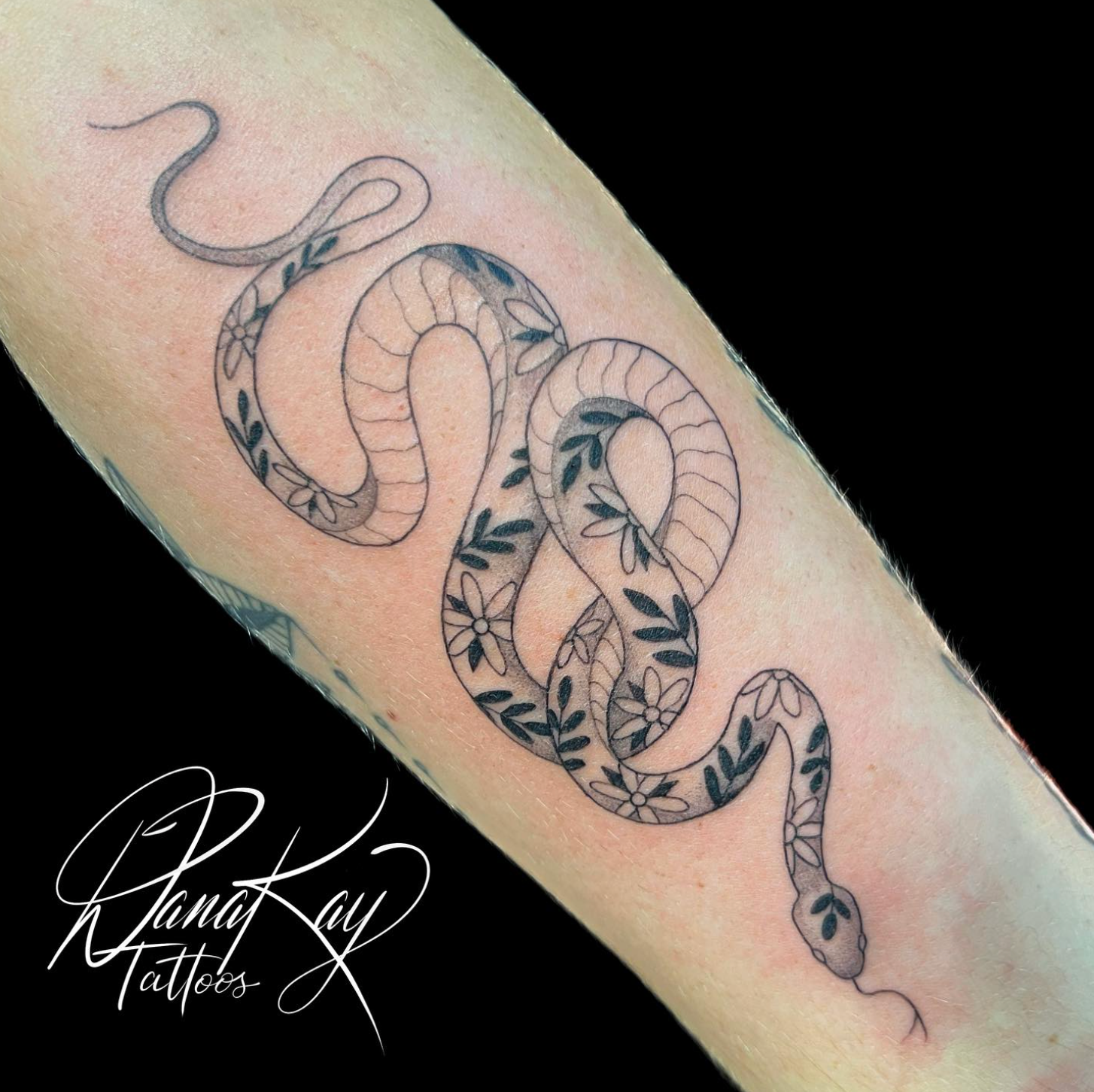 Tattoo of a snake with floral patterns on a person's arm. Black ink on skin.