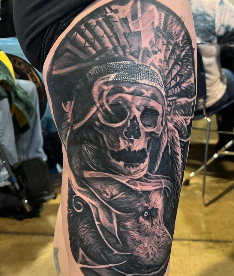 Skull wearing feathered headdress, tattoo on leg, with wolf-like figure.
