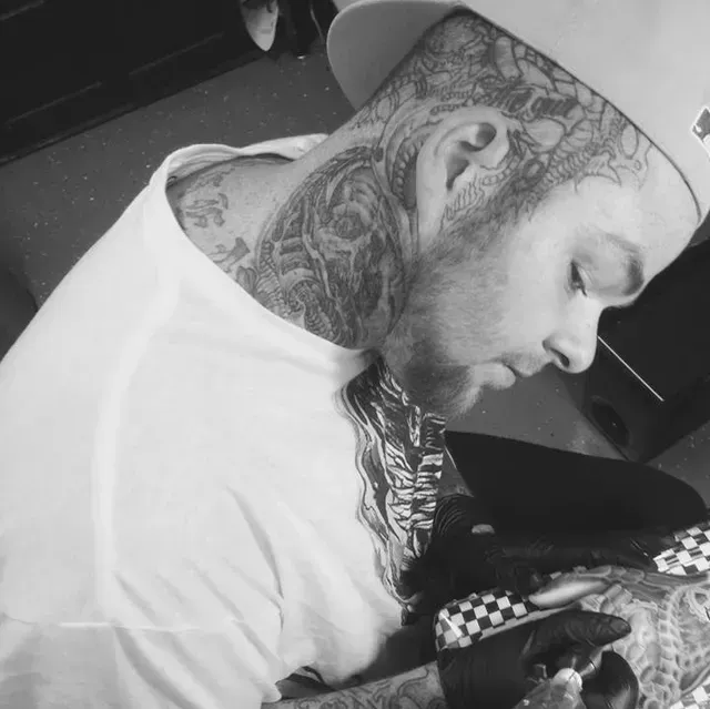 Man with neck tattoos, wearing a cap, looking down while working with gloved hands.