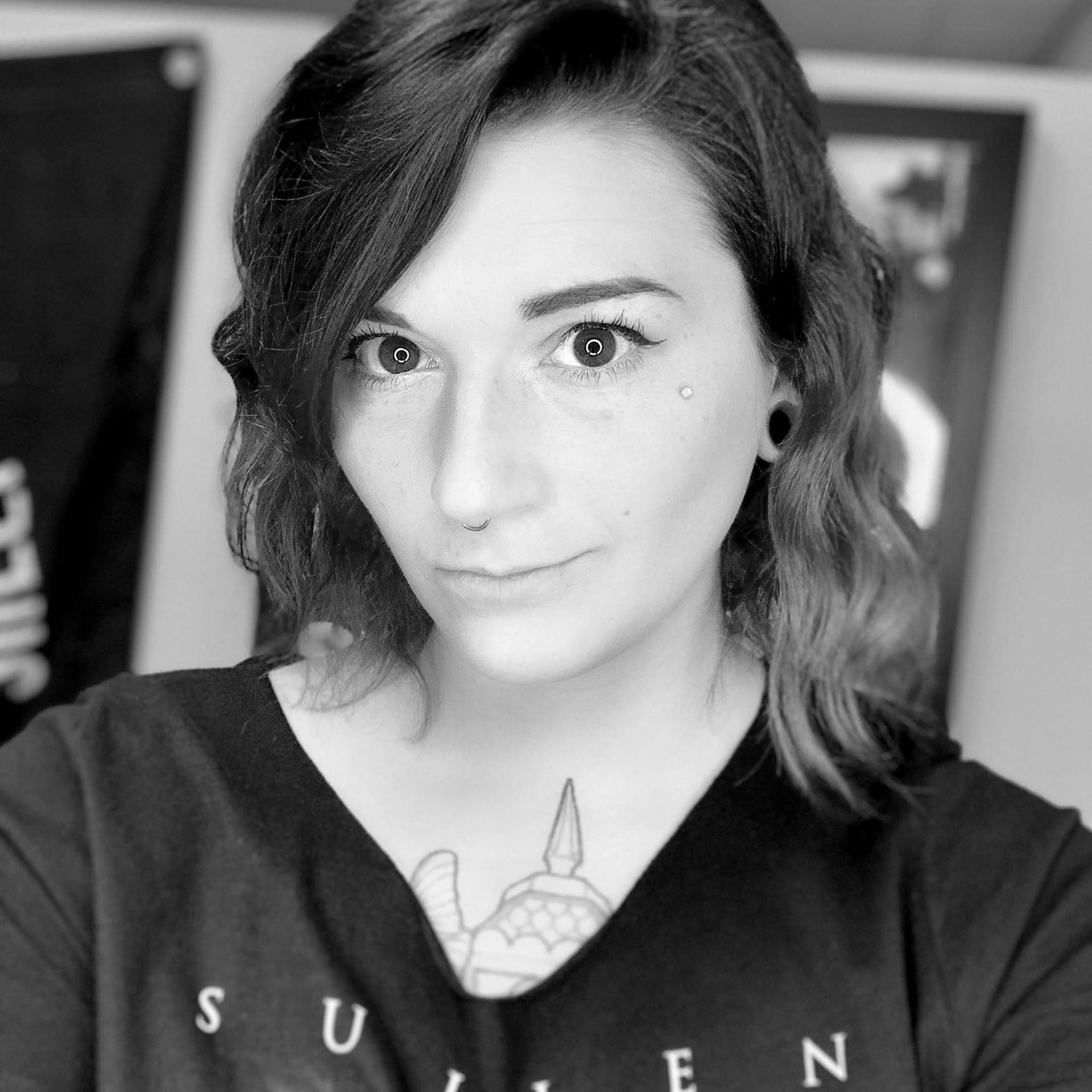Woman with wavy dark hair, nose ring, and gauges, looking at the camera. She wears a black shirt and has a neck tattoo.