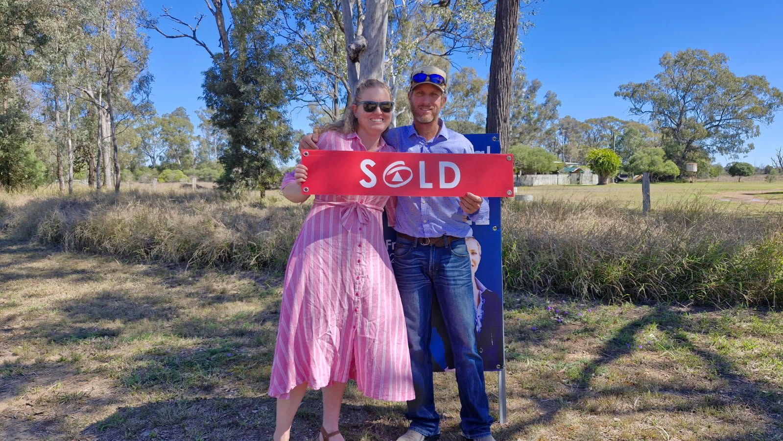 Couple holding a red “SOLD” sign in a sunny grassy field with trees behind them