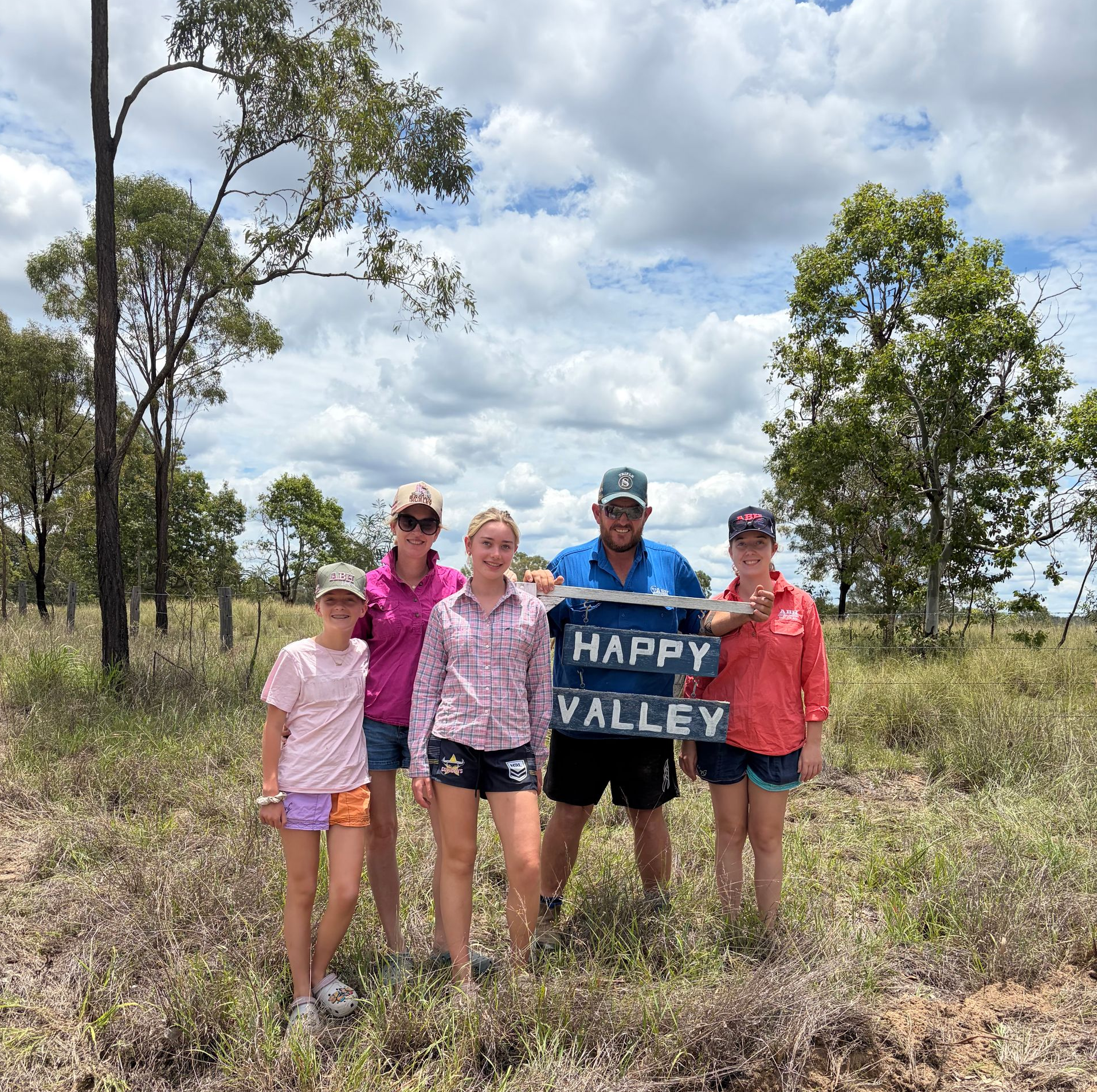 Five people standing in a grassy field holding a “HAPPY VALLEY” sign under a cloudy sky