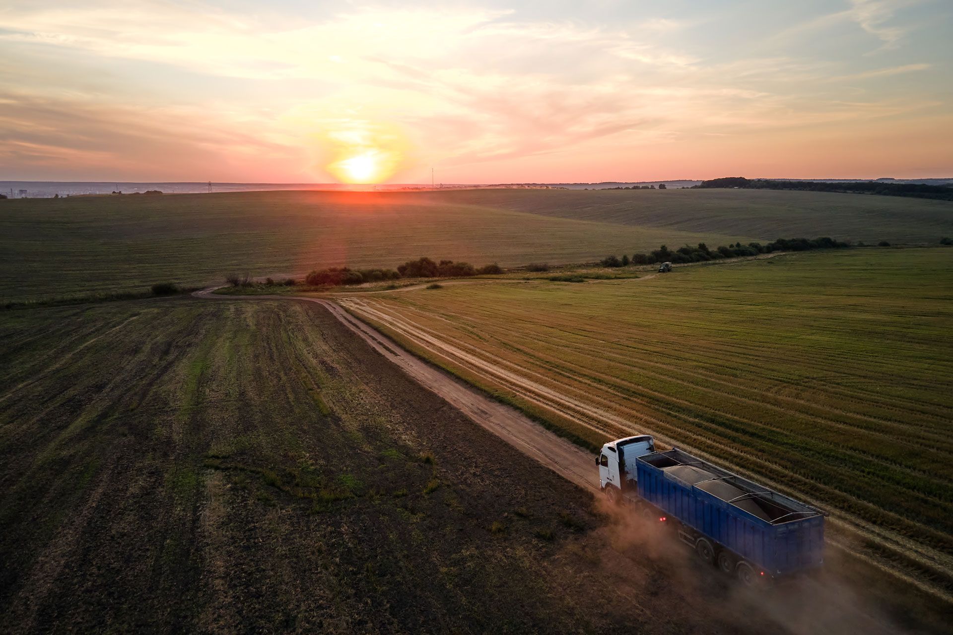 Tractor pulling a blue trailer along a dirt road through green fields at sunset
