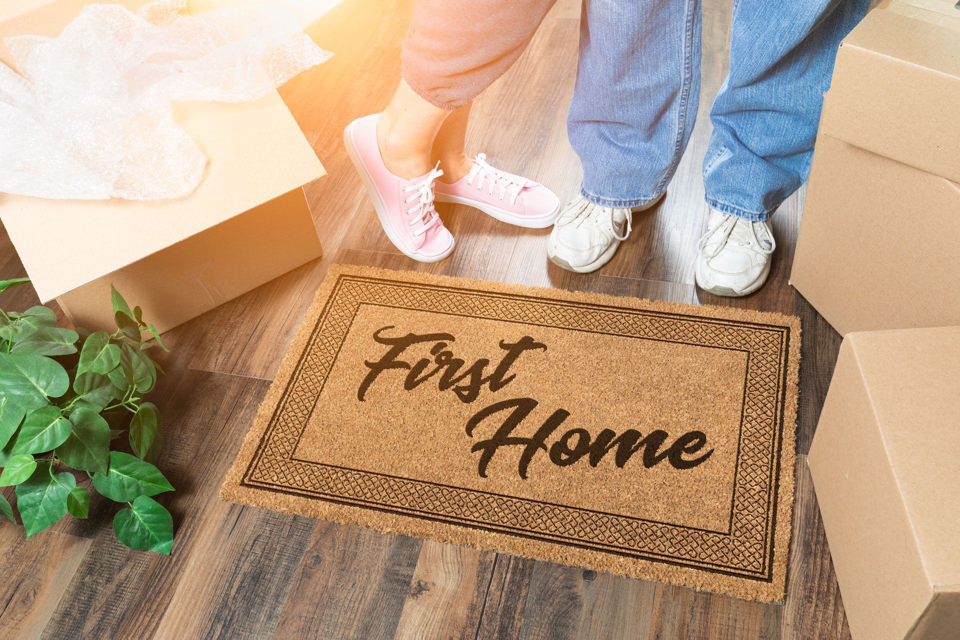 Couple’s feet beside a “First Home” doormat in a moving box-filled entryway