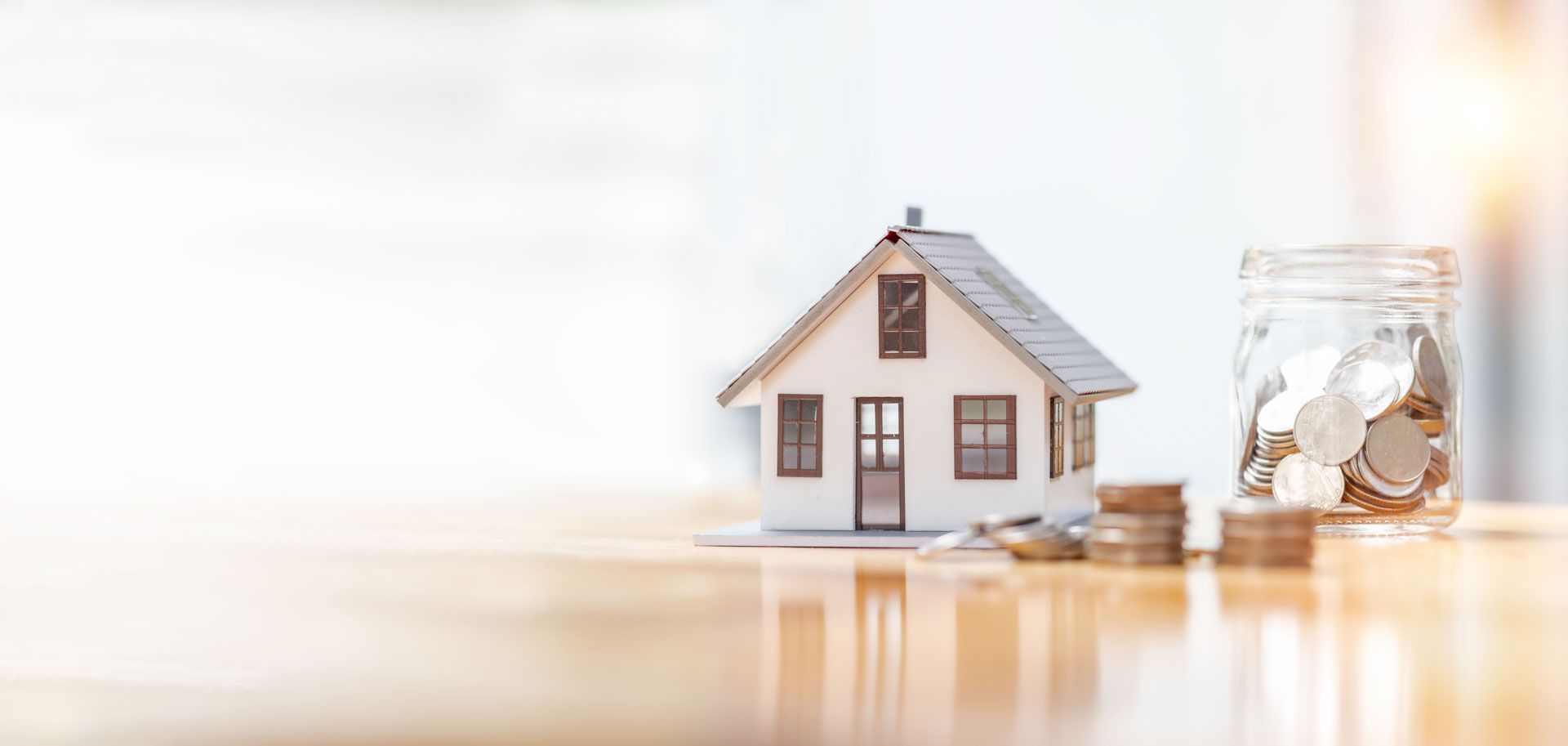 Miniature house model beside a glass jar of coins on a bright table, symbolizing home savings