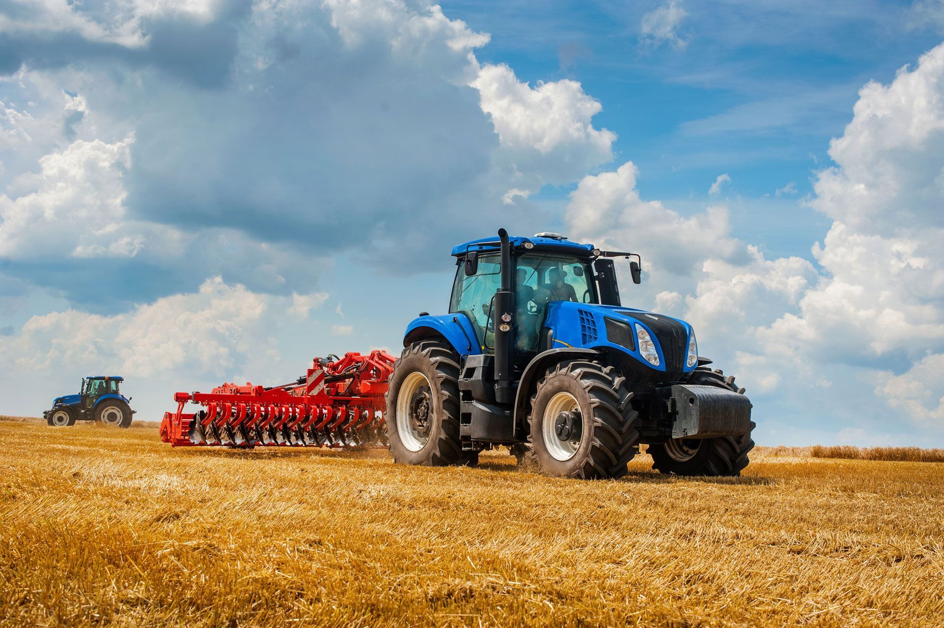 Blue tractor pulling red farm equipment across a golden field under a cloudy sky