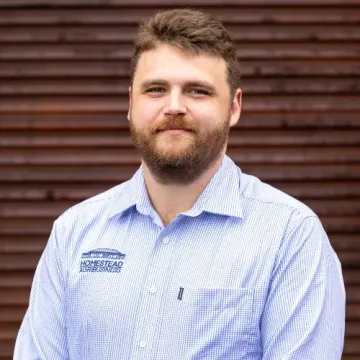 Domonic from Homestead Agribusiness ina light blue checkered shirt standing before a brown corrugated wall, smiling gently