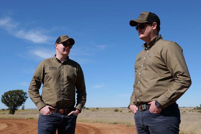 Two men in caps and sunglasses stand in a rural field under a clear blue sky.