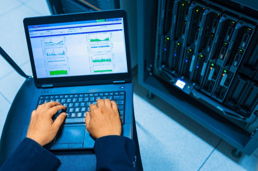 A person is typing on a laptop in a server room.