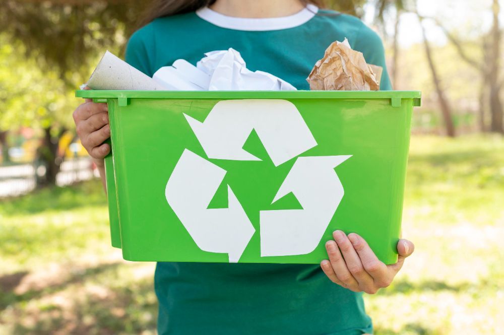 A woman is holding a green recycling bin filled with paper.