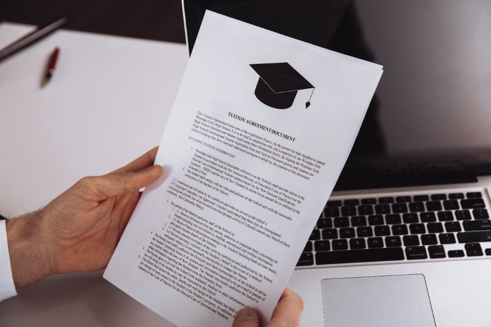 A person is holding a piece of paper with a graduation cap on it in front of a laptop.