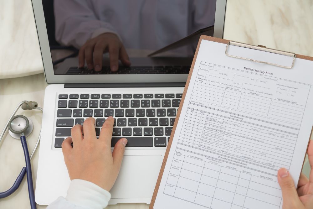 A doctor is typing on a laptop next to a clipboard and a stethoscope.