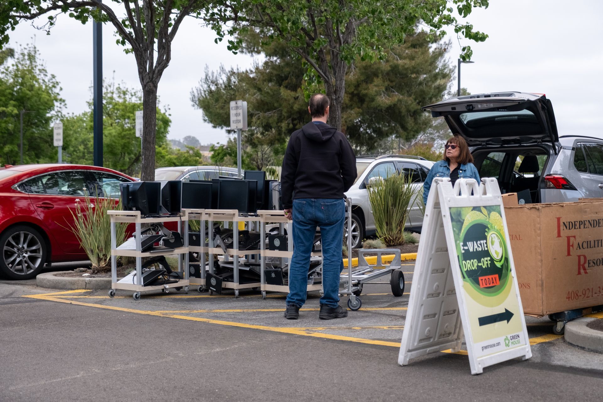 Man standing by tables of computer equipment in a parking lot, another person by open car trunk. An A-frame sign is nearby.