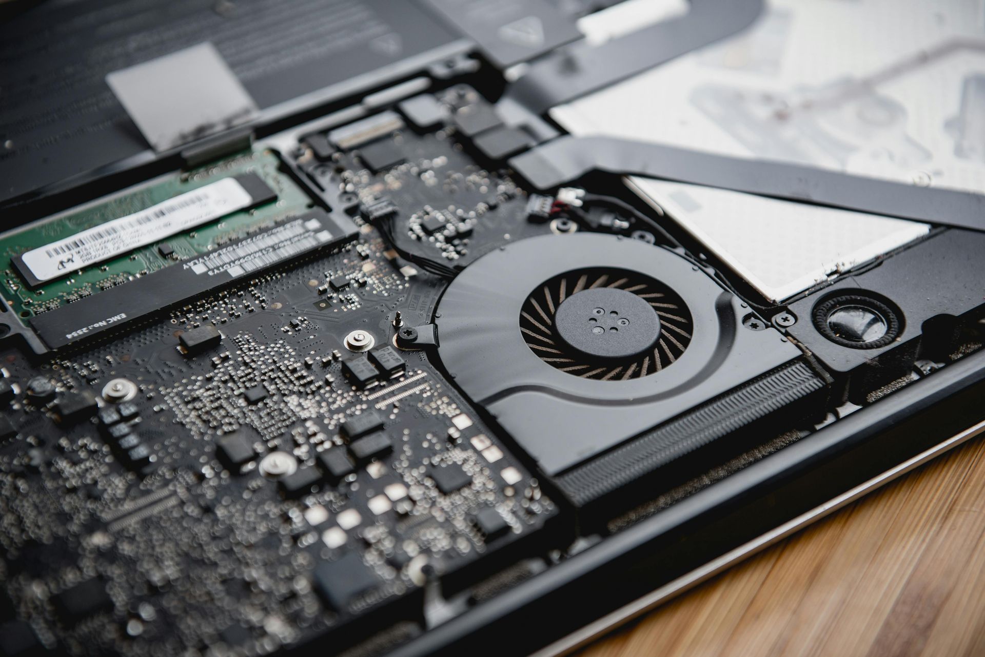 Laptop's internal components revealed: motherboard, RAM stick, fan, and circuitry on a wooden surface.