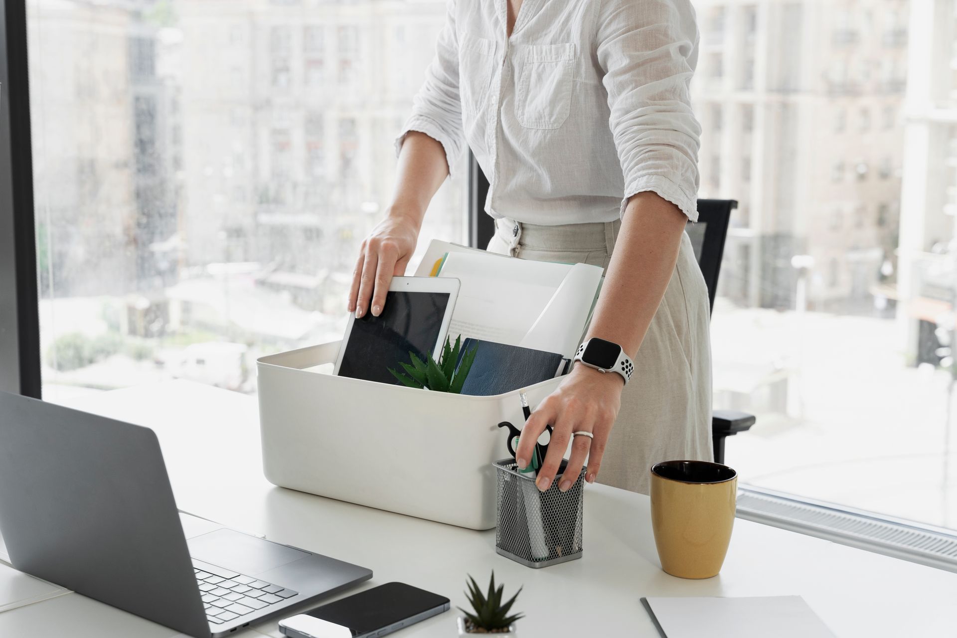 Person packing belongings into a box at an office desk. Sunlight streams through a large window.