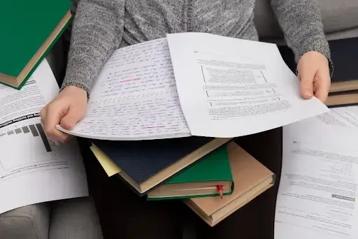 Person reviewing documents to shred in education, surrounded by books, handwritten notes, and printed academic materials.
