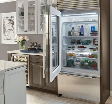 Open stainless steel refrigerator in a bright kitchen with shelves of food and drinks visible inside