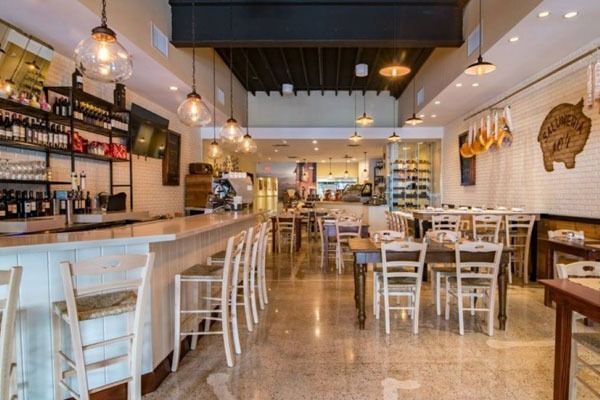 Bright, empty restaurant interior with white tables and chairs, wooden bar, and hanging pendant lights.