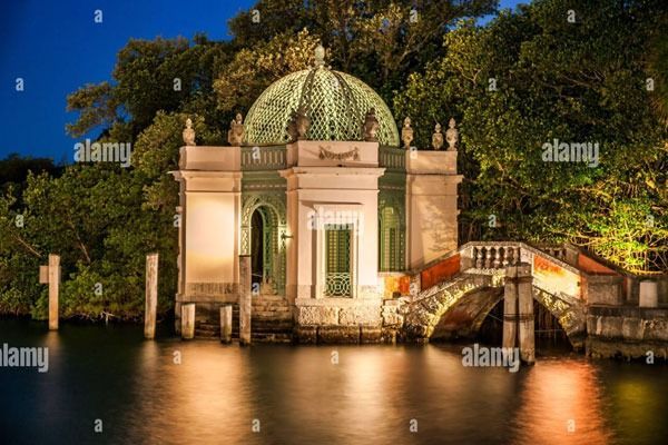 Illuminated domed pavilion by a river at night, with a small arched bridge and reflections on the water