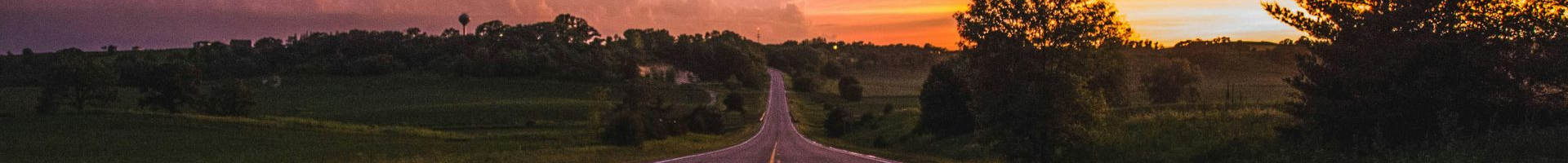 A road leads toward trees as the sun sets with hues of orange and purple.