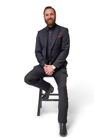 Man in dark suit sits on stool, smiling, hands clasped. Red pocket square. Black boots. White background.