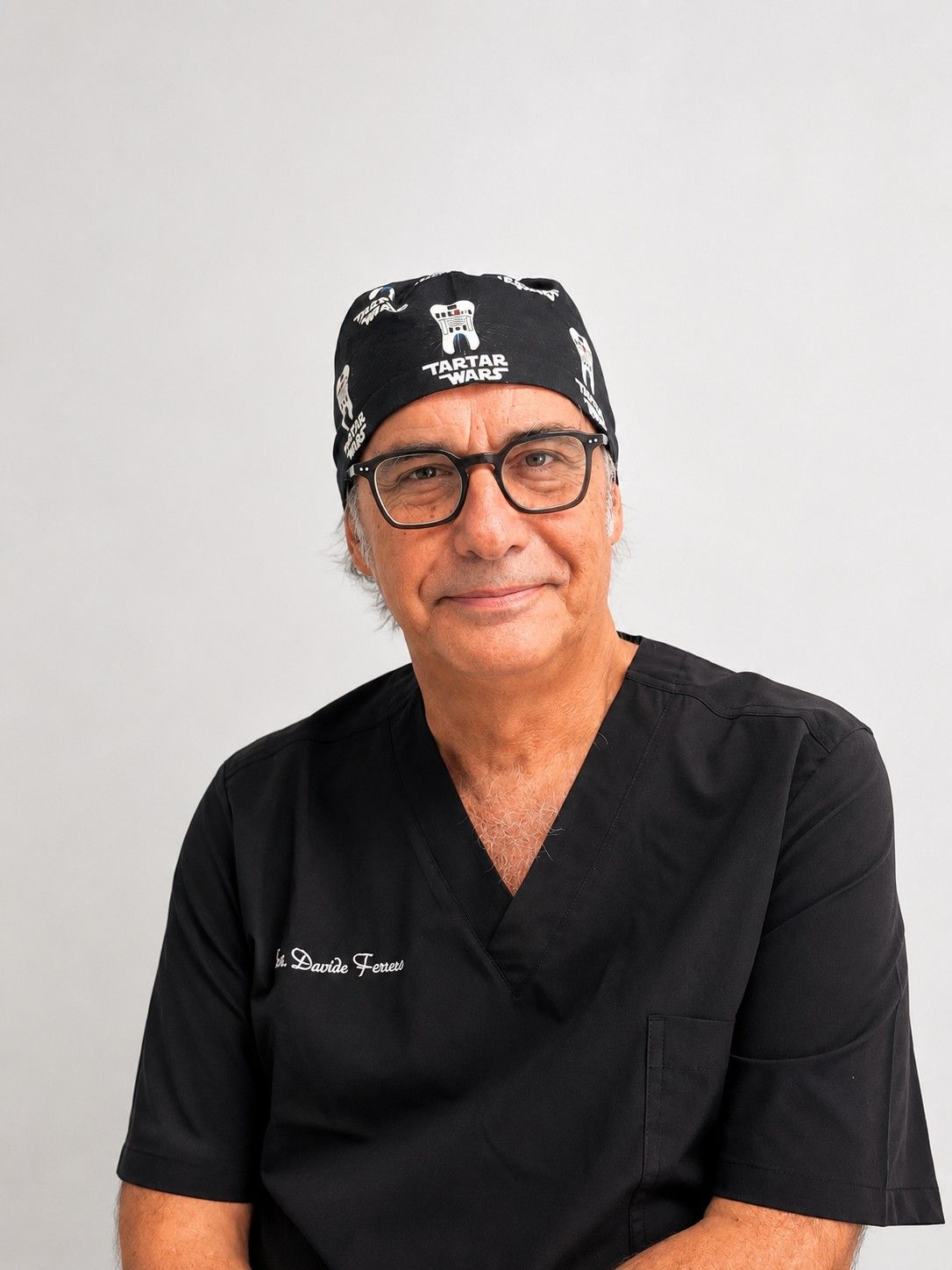 Ritratto di una persona sorridente in camice nero e bandana, su uno sfondo chiaro e uniforme.