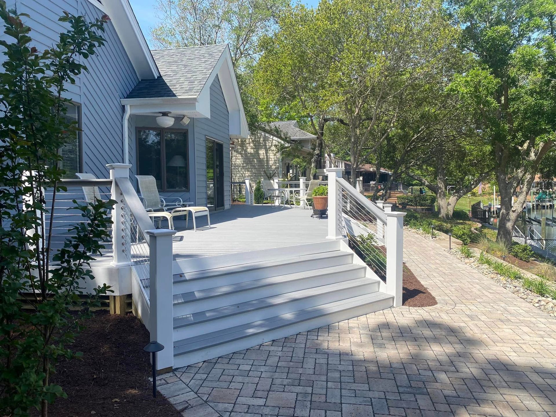 White front porch with steps and railing beside a blue house, lined by a paver walkway and trees