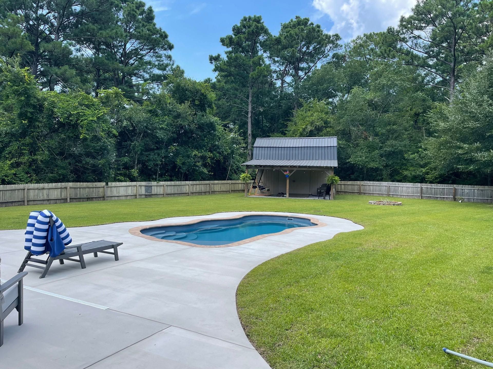 Backyard pool with blue lounge chair, concrete patio, and green lawn surrounded by trees