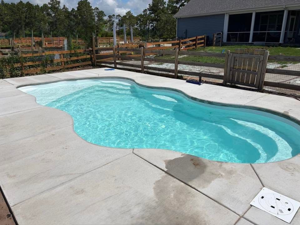 Backyard swimming pool with curved blue water, concrete deck, and wooden fence near a house