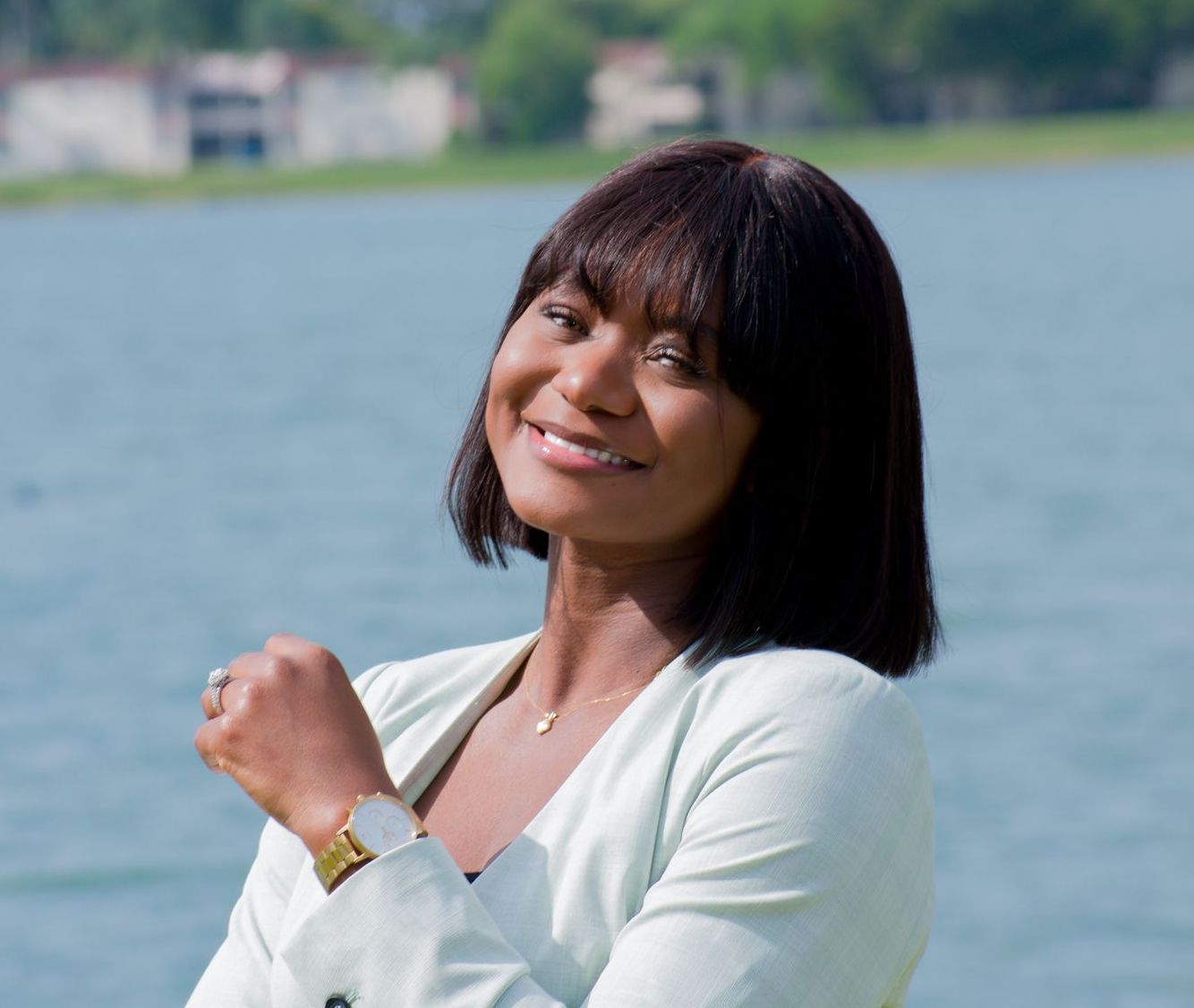 Smiling person by a lake, wearing a light cardigan and wristwatch, with blurred buildings in the background