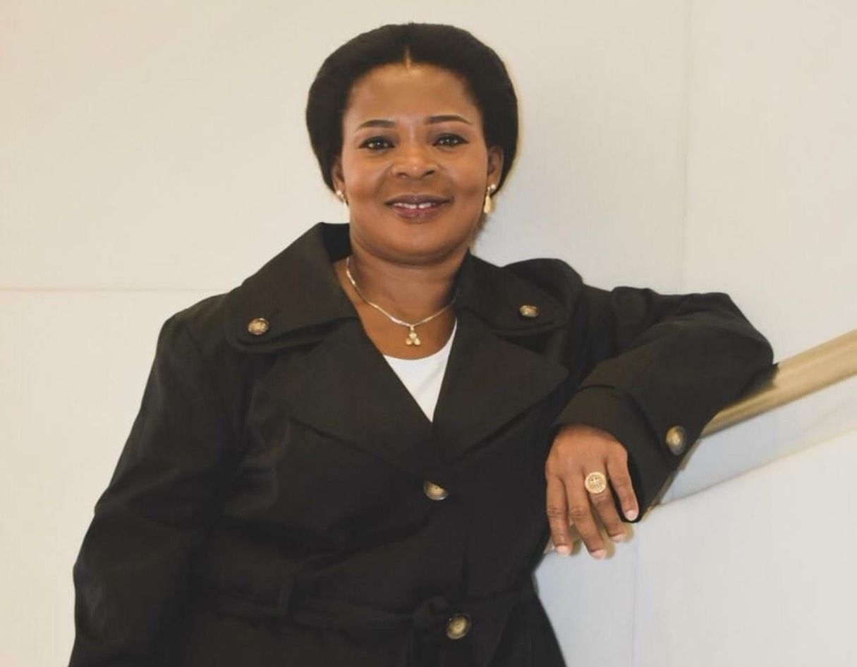 Smiling woman in a black suit leaning on a stair rail against a white wall