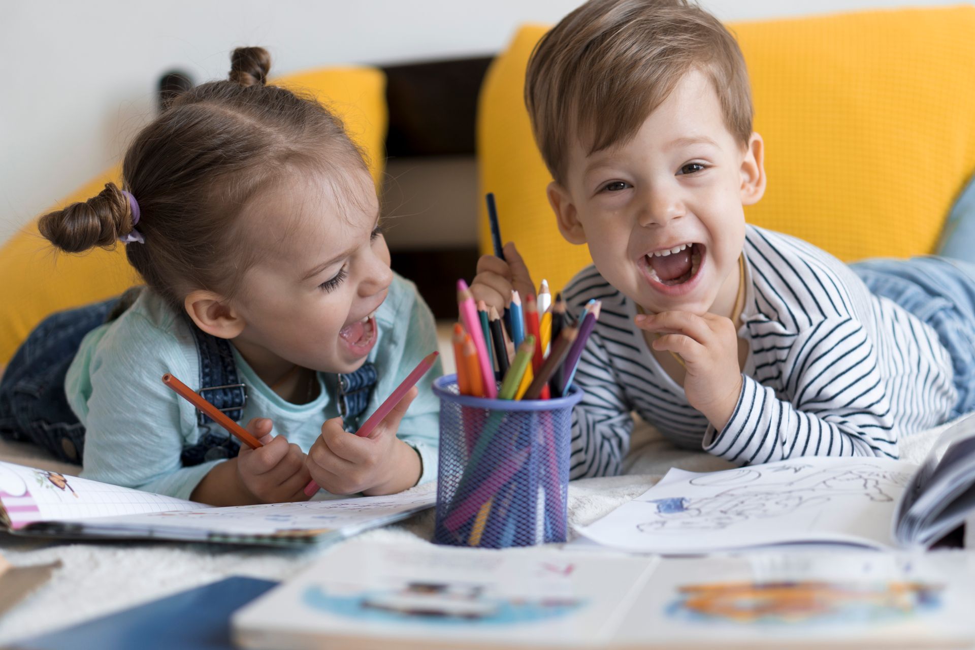 Two children drawing at a table with colored pencils, smiling in a bright room